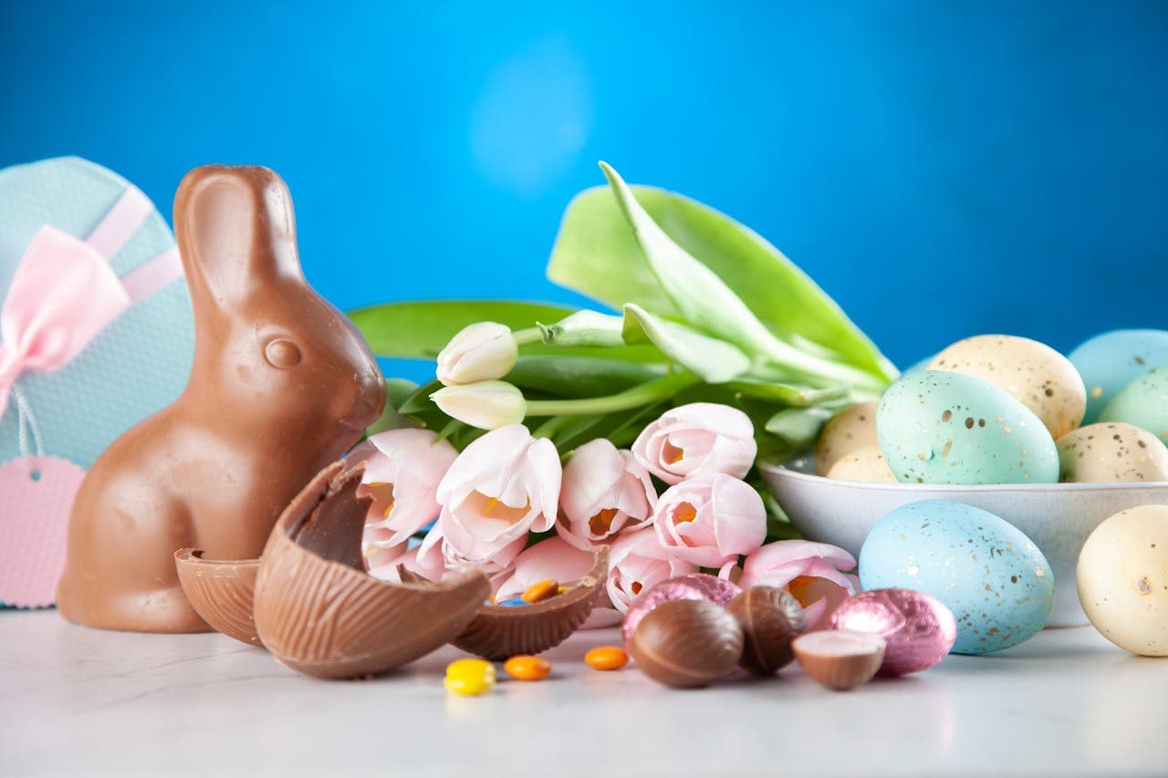 Festive Easter 2025 arrangement with chocolate bunny, pastel-colored Easter eggs, pink tulips, and sweet treats on a bright blue background – perfect for celebrating Easter traditions in Italy.