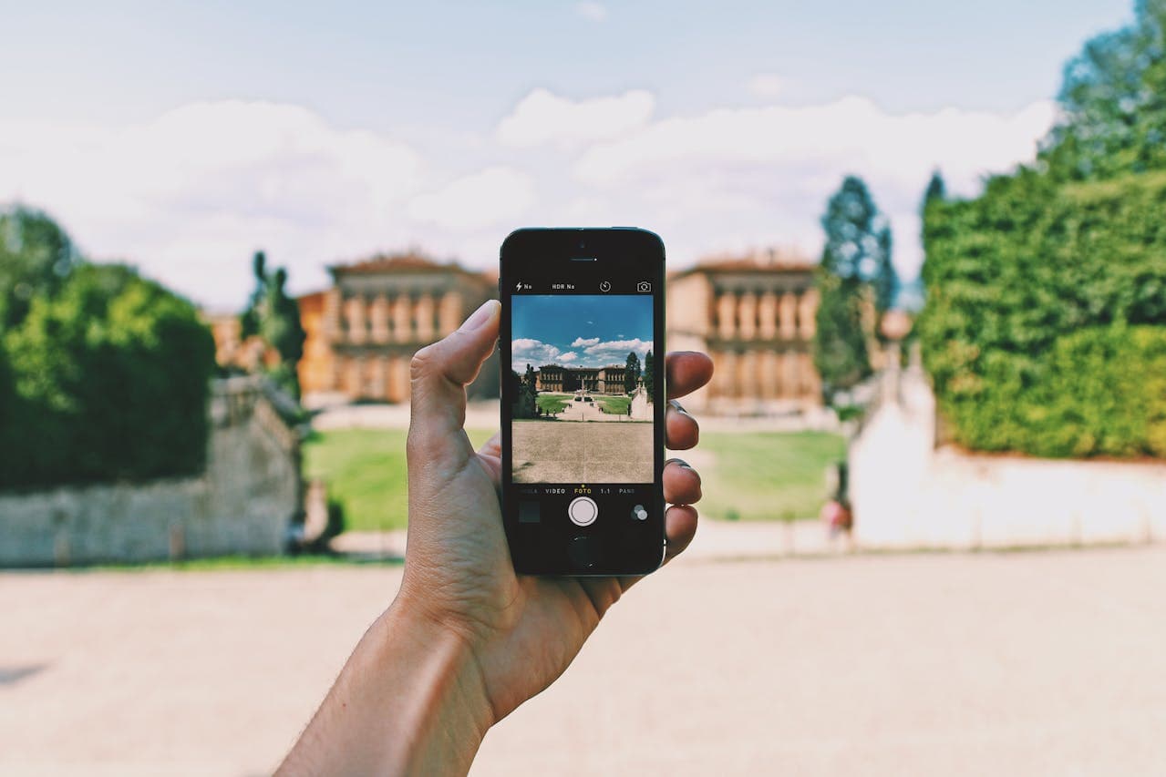 Hand holding smartphone taking a photo of the Boboli Gardens in Florence, Italy, during a guided tour with Palazzo Pitti in the background.