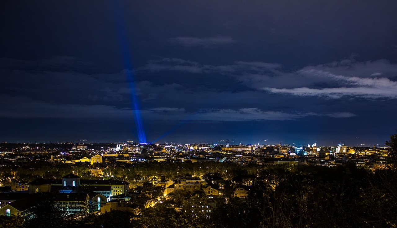 Panoramic view of Rome at night from Gianicolo Hill with city lights and blue spotlights illuminating the sky.