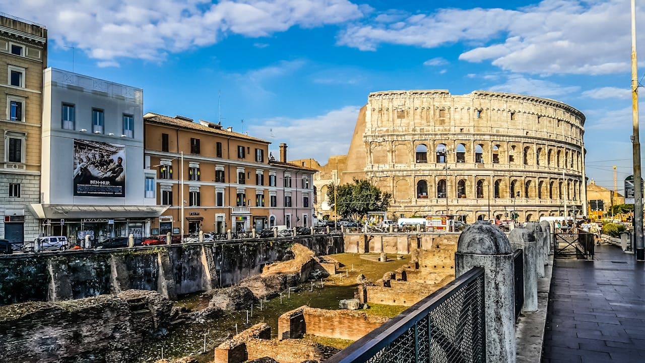 View of the Colosseum in Rome with nearby ruins and modern buildings under a blue sky — perfect starting point for a Colosseum tour.
